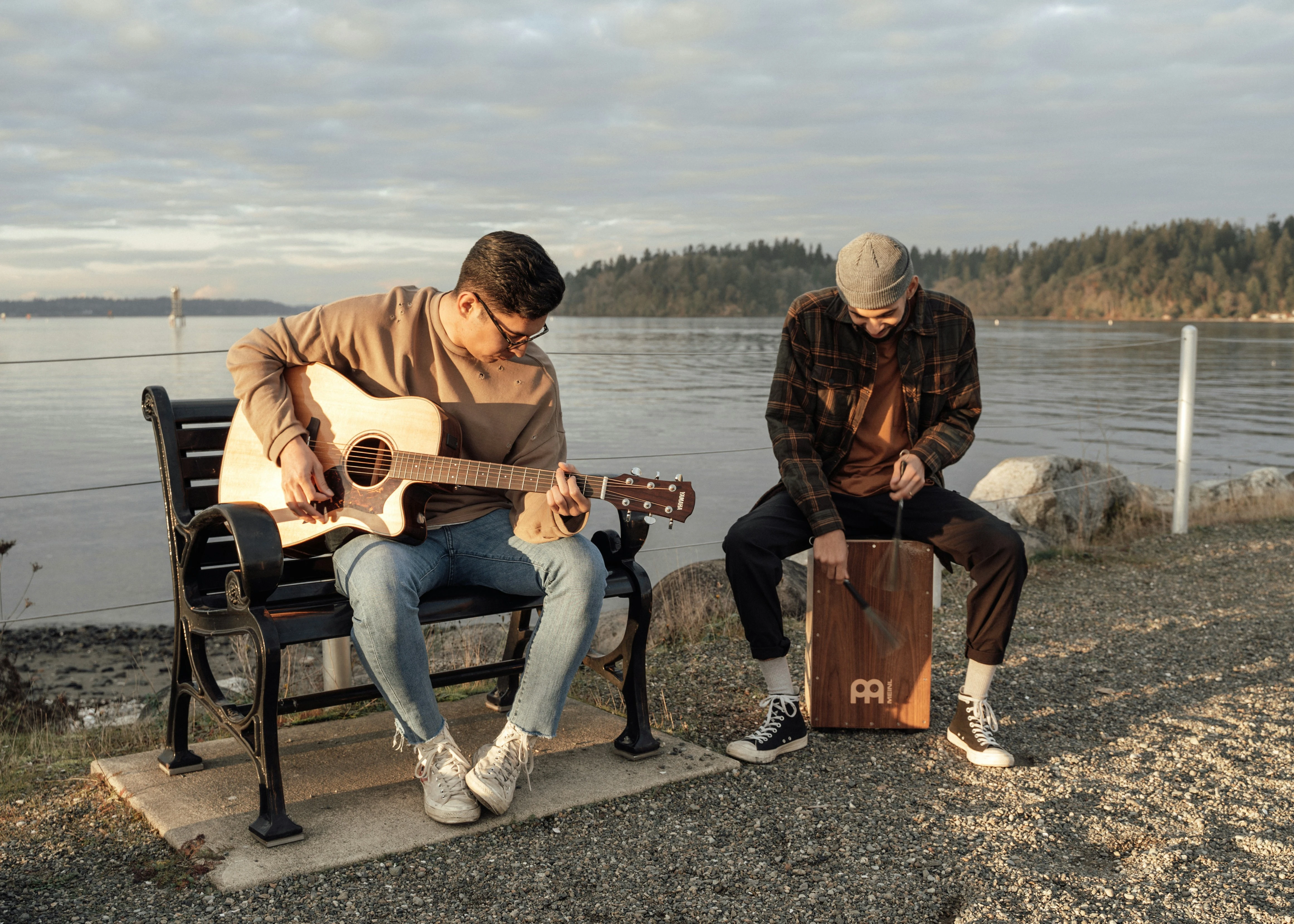two men are sitting on a bench, one is playing guitar, the other sitting on a drum box that he is playing